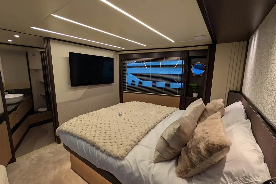 Modern yacht bedroom featuring a plush bed, wall-mounted TV, elegant light fixtures, and a window with blinds.